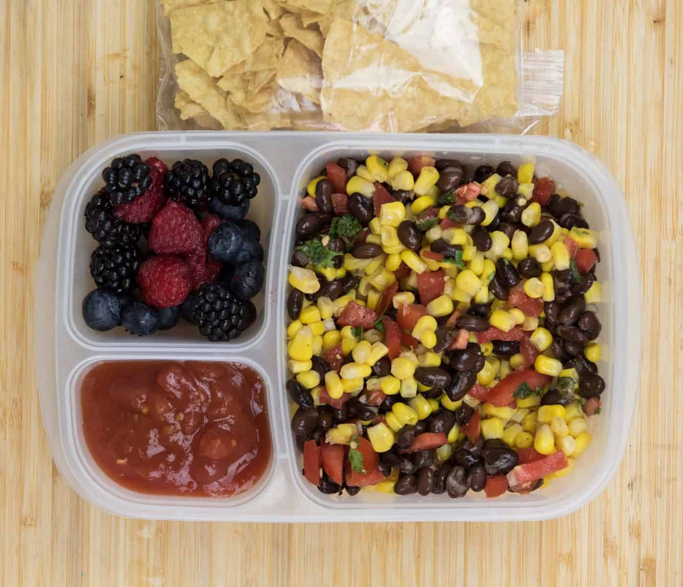 Top view of a lunch box with Corn and Bean Salad, Salsa, Tortilla chips, and Mixed berries.