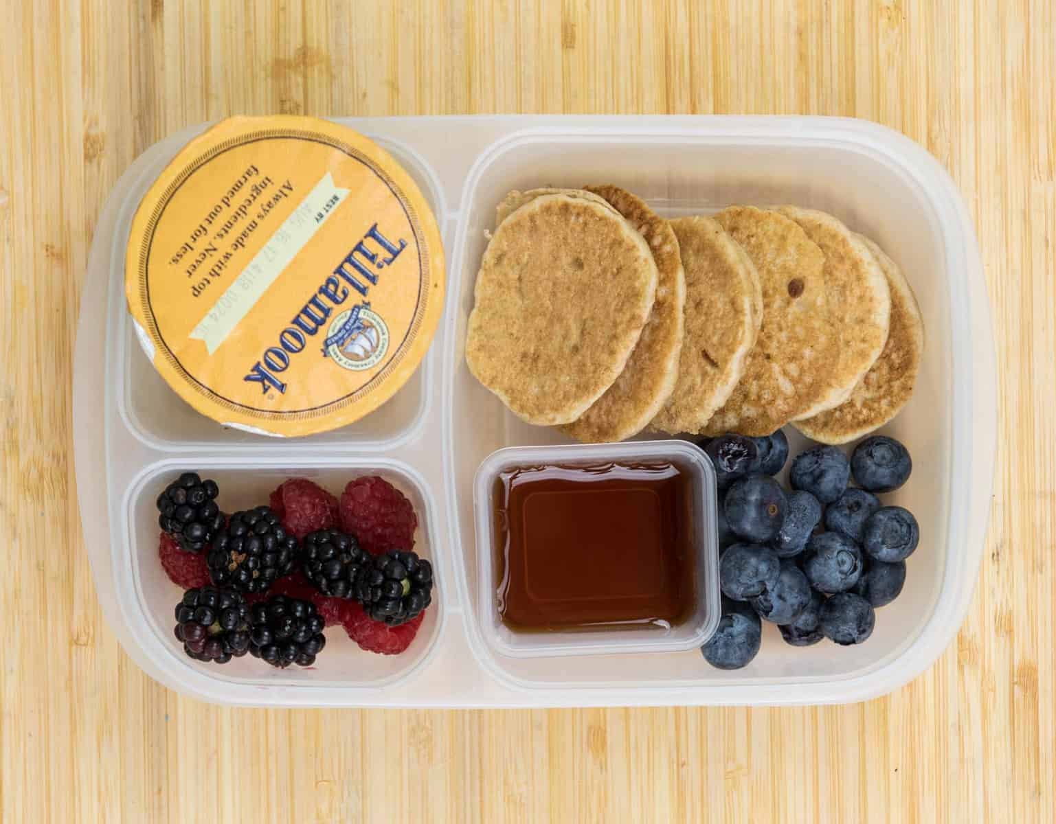 Top view of a lunch box with Mini whole wheat pancakes, Maple syrup, Blackberries + raspberries + blueberries, and Yogurt.