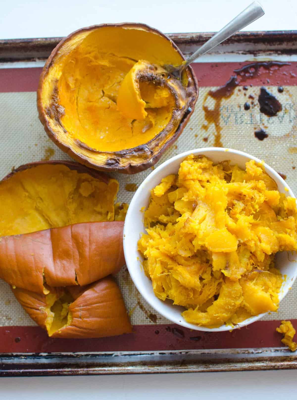 Cooked pumpkin in a white bowl next to a cooked pumpkin half with a spoon and an empty pumpkin shell on a rimmed baking sheet lined with a silicone baking mat.