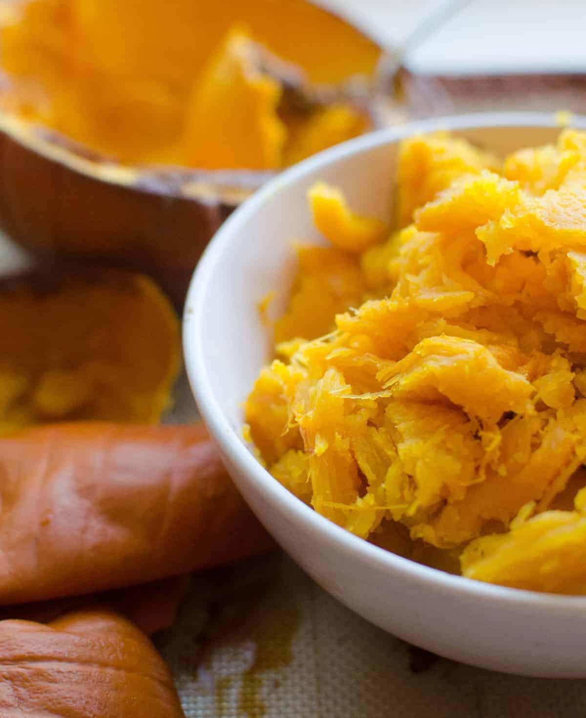 Cooked pumpkin in a white bowl next to an empty cooked pumpkin shell.