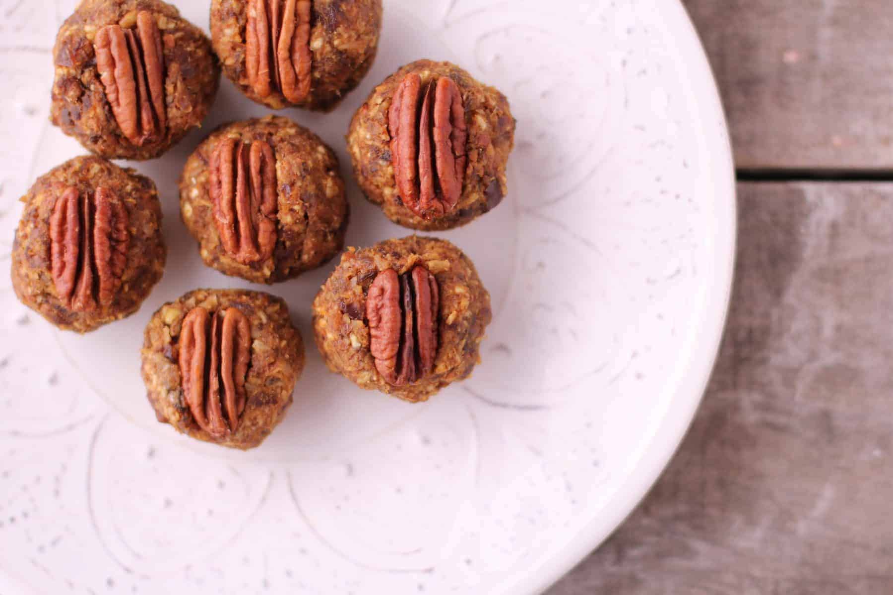 A white plate with seven pumpkin date balls topped with pecan halves, arranged on a rustic wooden surface. The snacks boast a textured, nutty exterior.