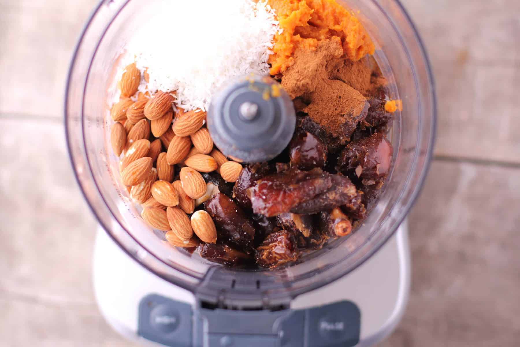 A food processor filled with whole almonds, shredded coconut, chopped dates, cinnamon, and a scoop of sweet potato whirls into a mix perfect for pumpkin date balls.