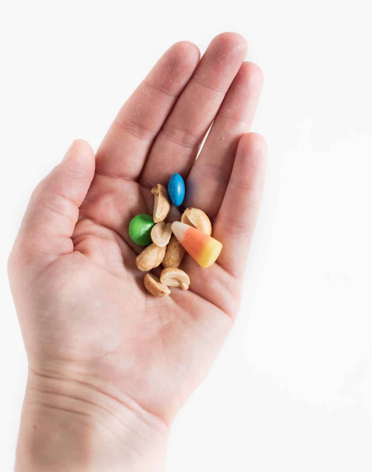 A hand holding some candy corn snack mix made with M&Ms, roasted peanuts, and candy corn in a blue bowl.