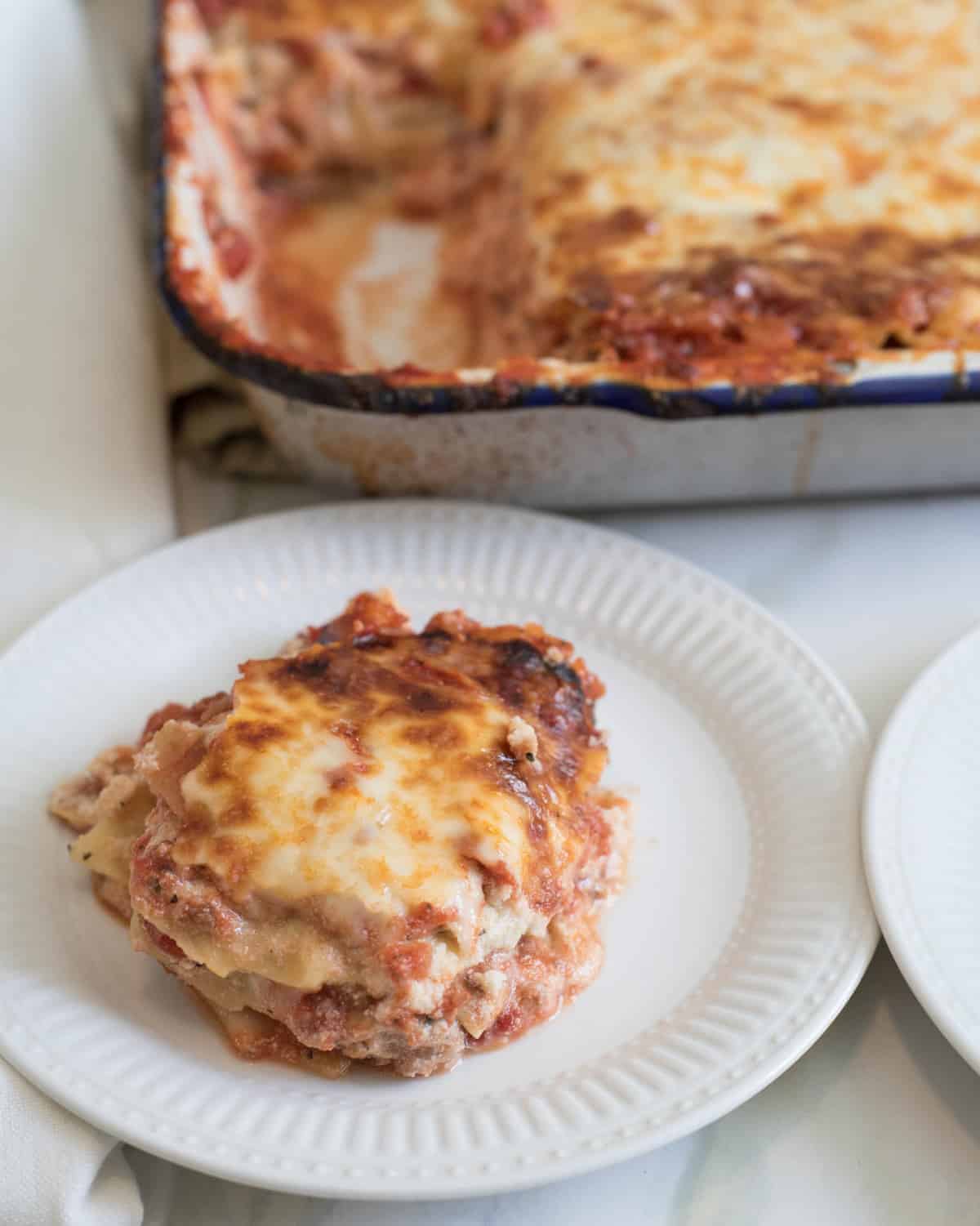 A serving of baked lasagna recipe with melted cheese on top sits on a white plate in the foreground, with a tray of the remaining lasagna in the background.