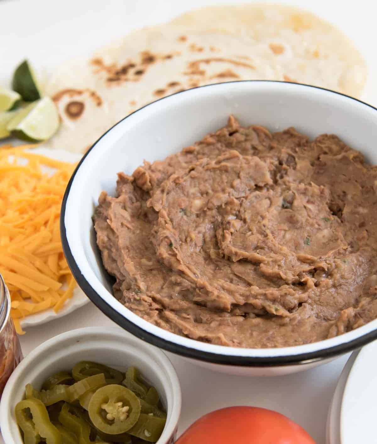 A close-up of refried beans in a white enamel bowl surrounded by taco fixings, including tortillas, shredded lettuce, shredded cheddar cheese, salsa, sliced jalapeños and a tomato.
