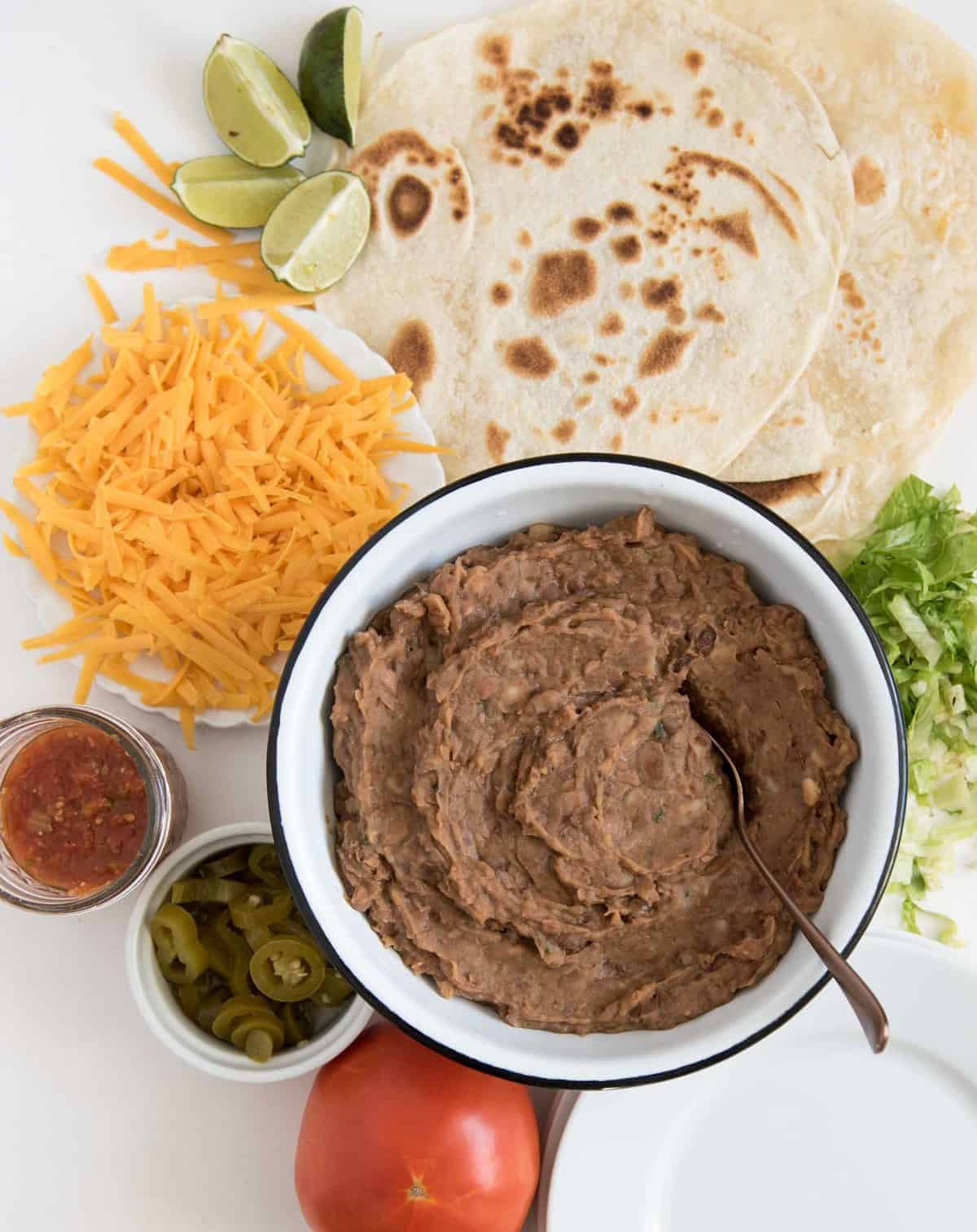 Refried beans in a white enamel bowl with a spoon in it surrounded by taco fixings, including tortillas, shredded lettuce, shredded cheddar cheese, salsa, sliced jalapeños and a tomato.