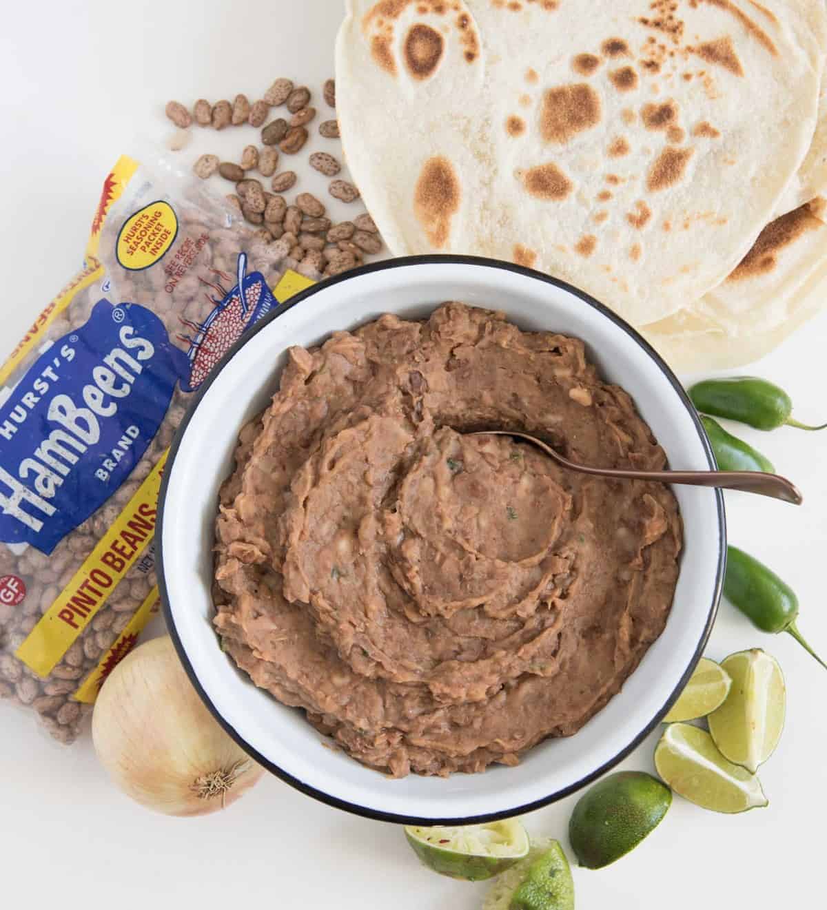 Refried beans in a white bowl with spoon in it and a bag of pinto beans, onion, some lime slices, tortillas, and whole jalapeños next to it.