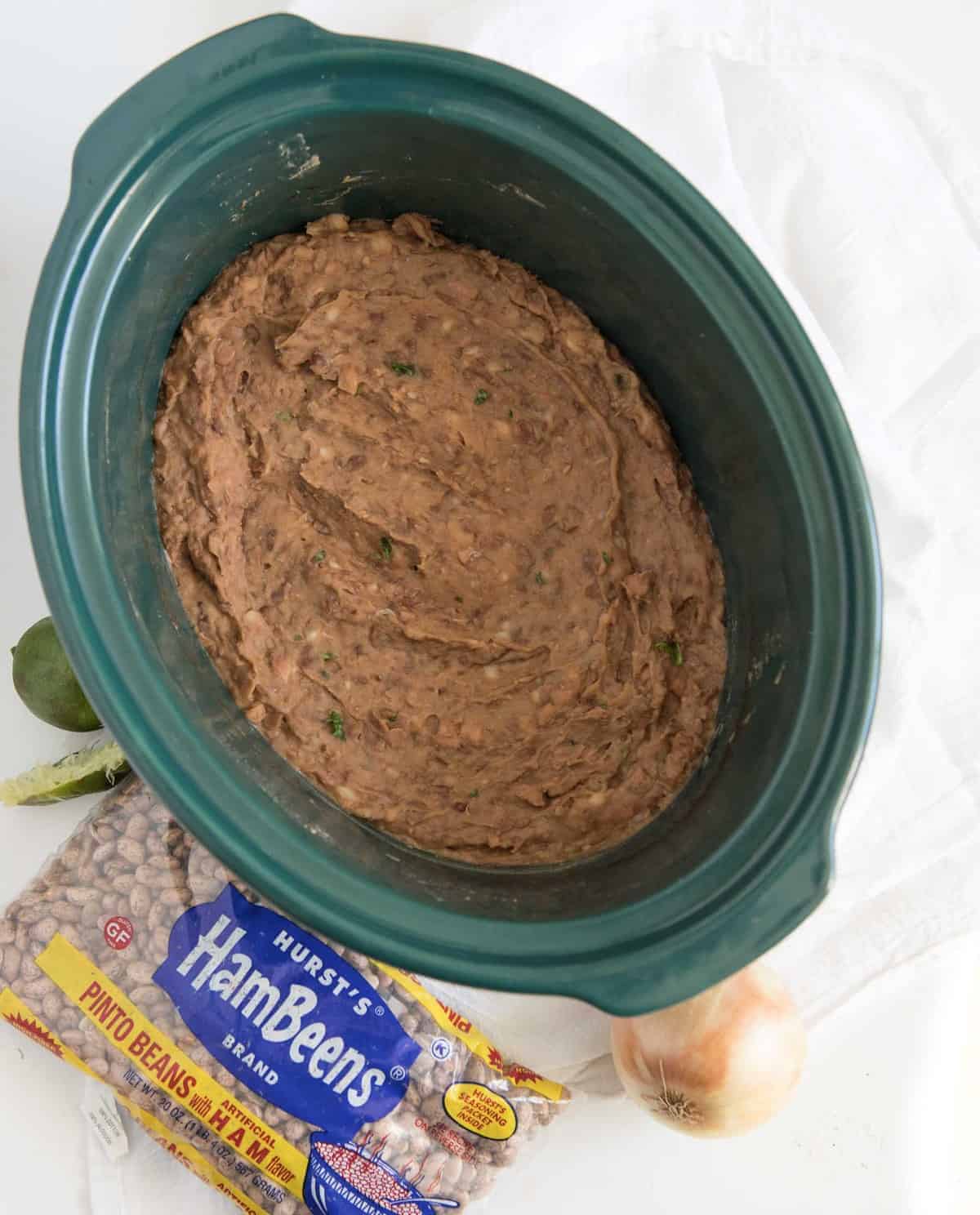 Refried beans in a dark green slow cooker with a bag of pinto beans, an onion and a lime next to it.