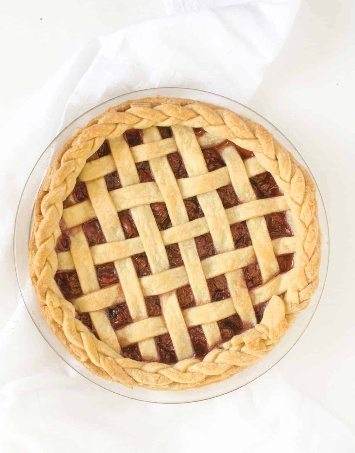A golden-brown cherry pie with a lattice crust and braided edge sits on a clear glass pie dish atop a white cloth, reminiscent of the classic charm found in a homemade coconut cream pie.