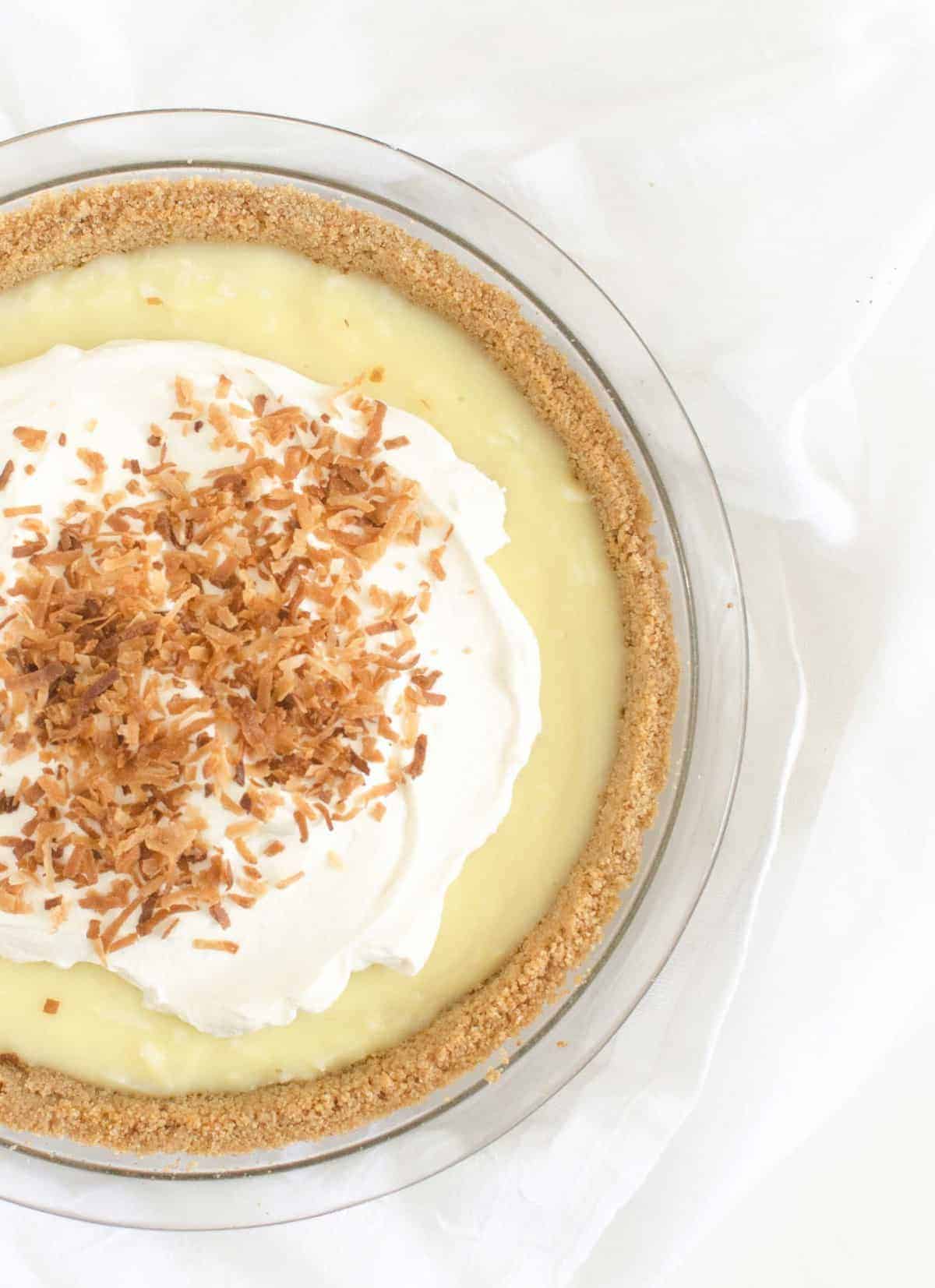 A classic coconut cream pie sits in a glass dish, topped with fluffy whipped cream and toasted shredded coconut, all displayed on a white background.