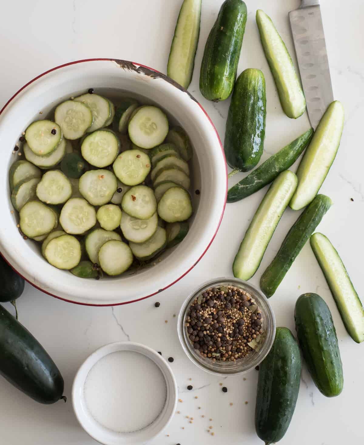 Top view of a bowl of cucumbers, slice cucumbers on the side, and a jar of pickling spice.