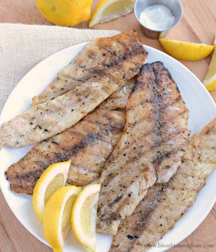 Grilled fish fillets seasoned with pepper rest on a white plate, served with lemon wedges. Perfect for grilling fish enthusiasts, this dish is accompanied by a small metal cup of salt and extra lemon slices on a light surface.