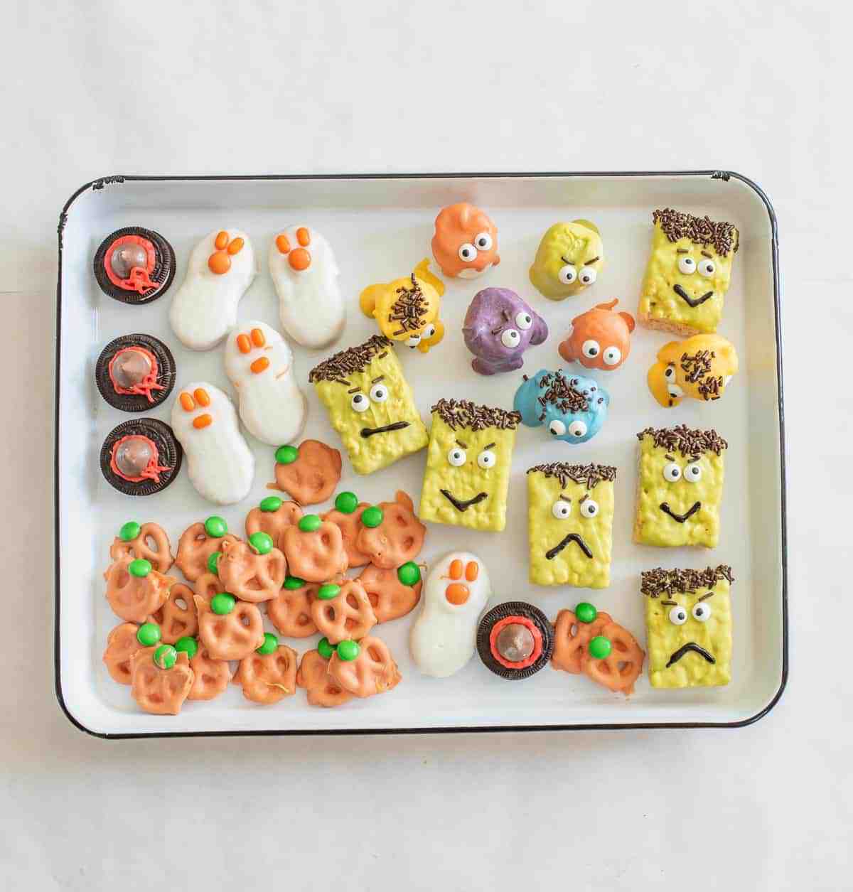 A tray of no bake Halloween treats featuring ghost-shaped cookies, Frankenstein square bites with sprinkles, monster-faced candies, and pretzels coated in orange with green candies for a festive touch.