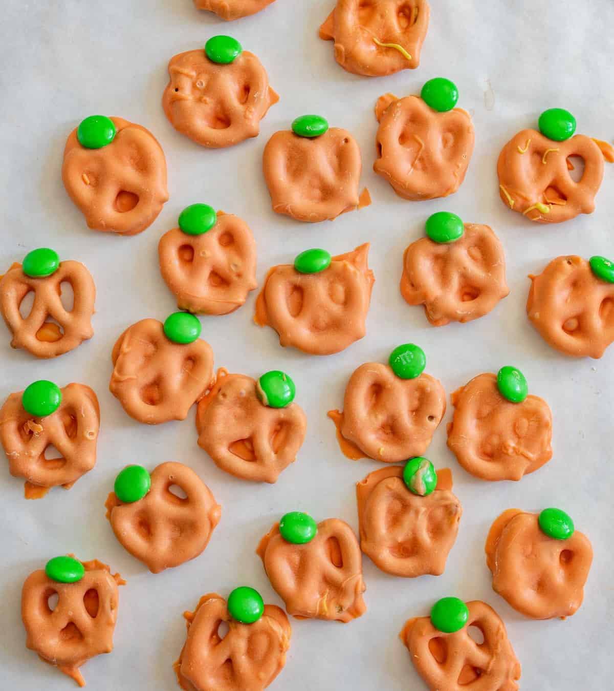Pretzels coated in orange candy are arranged to resemble pumpkins, each topped with a green candy for the stem—perfect as festive treats or creative lunch box ideas, all displayed on parchment paper.
