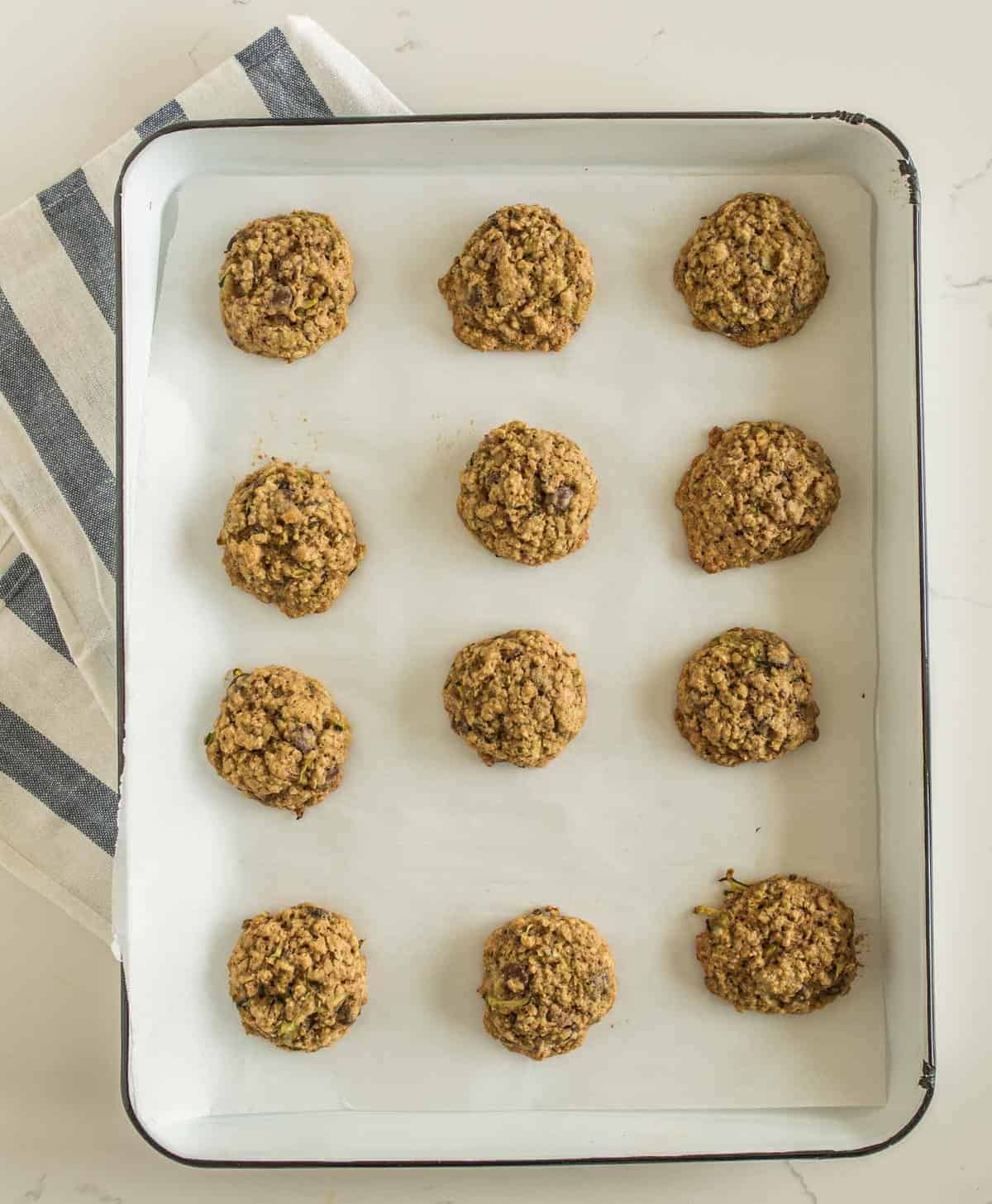 A baking tray lined with parchment paper holds twelve evenly spaced oatmeal cookies with a rustic texture, resembling the hearty feel of taco zucchini boats. The tray rests on a striped cloth on a light countertop.