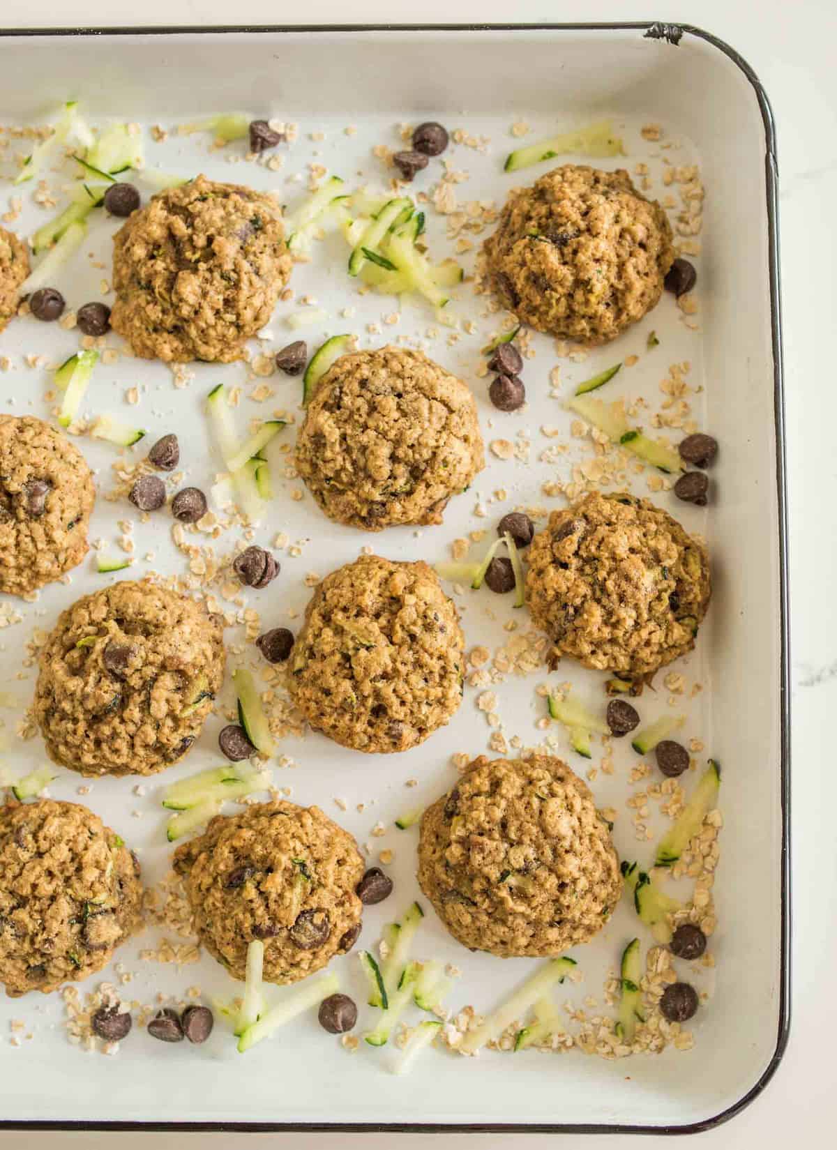 A white baking tray showcases ten delightful zucchini cookies with oatmeal, each scattered with chocolate chips and shredded zucchini. Arranged in three neat rows, oats and zucchini are artfully sprinkled around to enhance their rustic charm.
