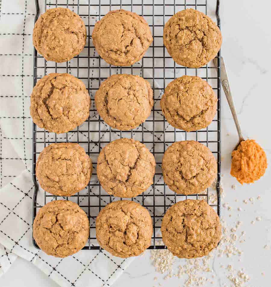 Twelve pumpkin spice muffins cooling on a wire rack, with a spoonful of orange batter beside them and a white cloth with a black grid pattern underneath. Some oats are scattered nearby on the surface.