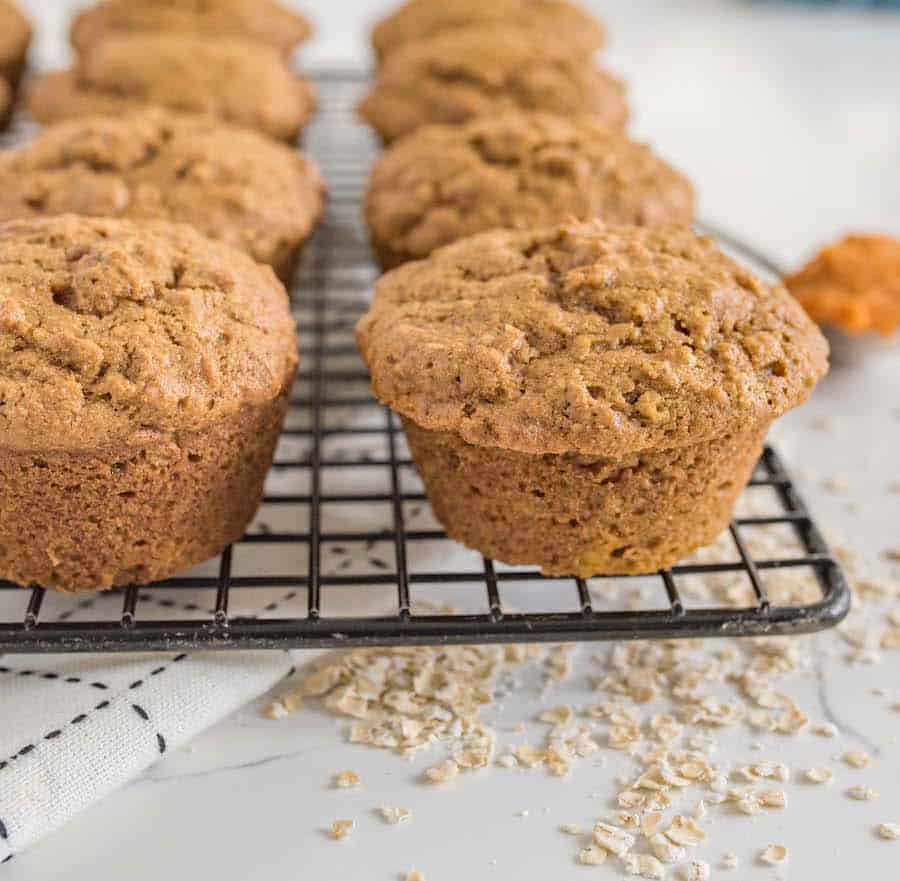 A close-up of several freshly baked pumpkin spice muffins on a cooling rack, with oats scattered on a white surface and a napkin partially visible underneath.