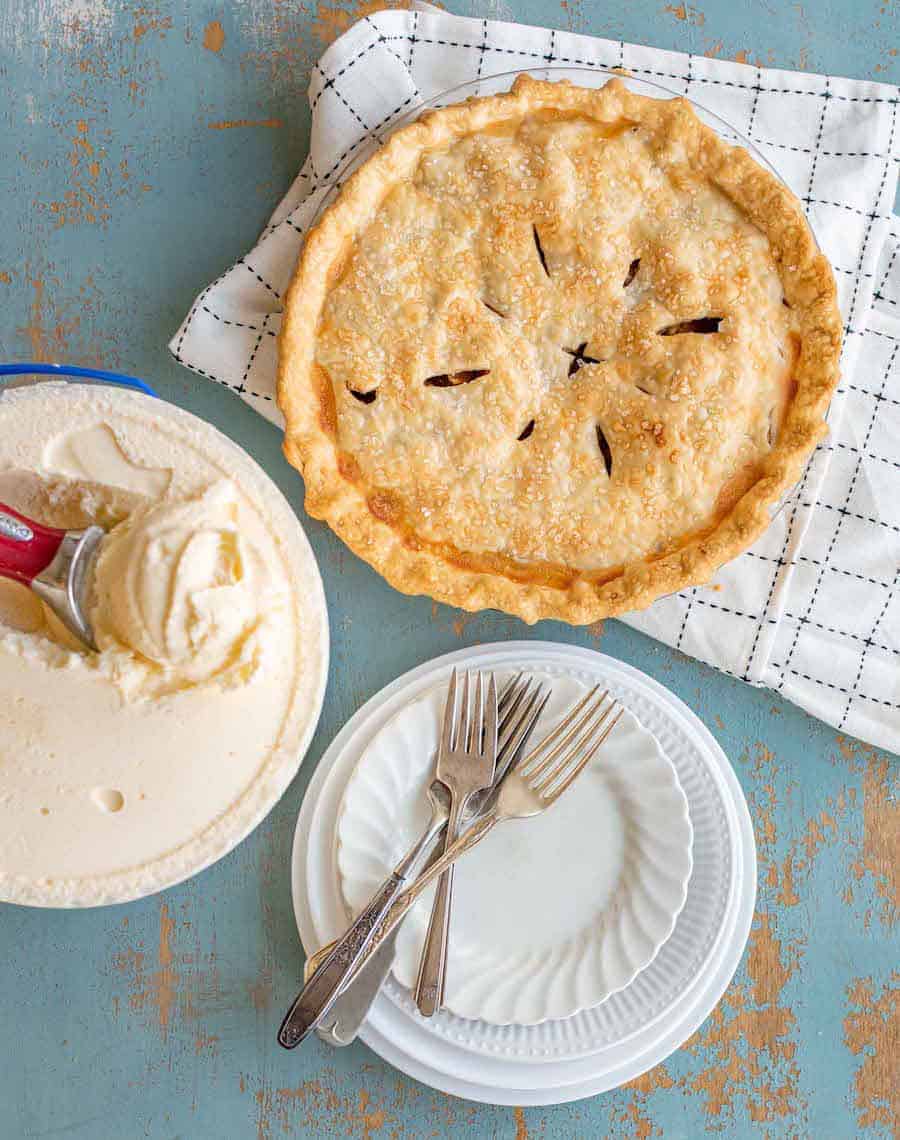 An apple pie made from a simple apple pie recipe rests on a checkered cloth beside vanilla ice cream with a scooper, and a stack of plates with forks on a blue rustic surface.