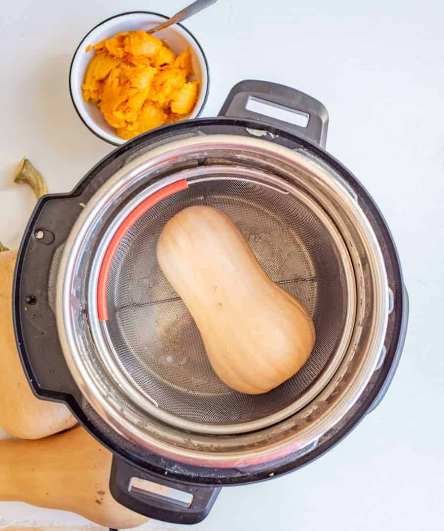 Image of a whole butter nut squash sitting in a steam basket inside an Instant Pot with a bowl of cooked butternut squash in the background.