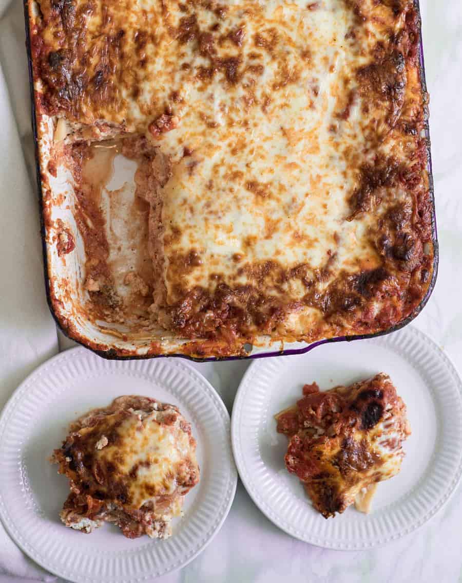 A large baked lasagna from an Easy Lasagna Recipe sits in a rectangular dish with a portion missing, beside two white plates, each holding a serving of the cheesy, golden-brown lasagna on a light-colored surface.