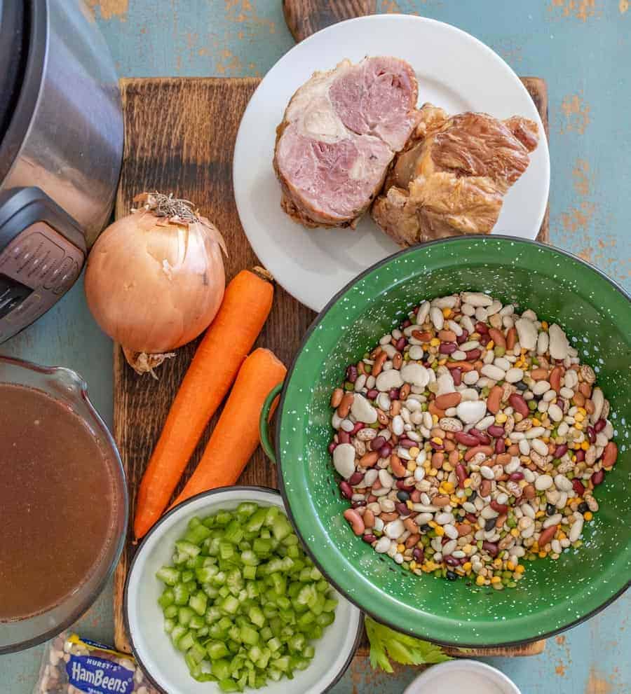 Top view of a green bowl of dried beans with white bowl filled with diced celery on cutting board with whole carrots yellow onion and ham.