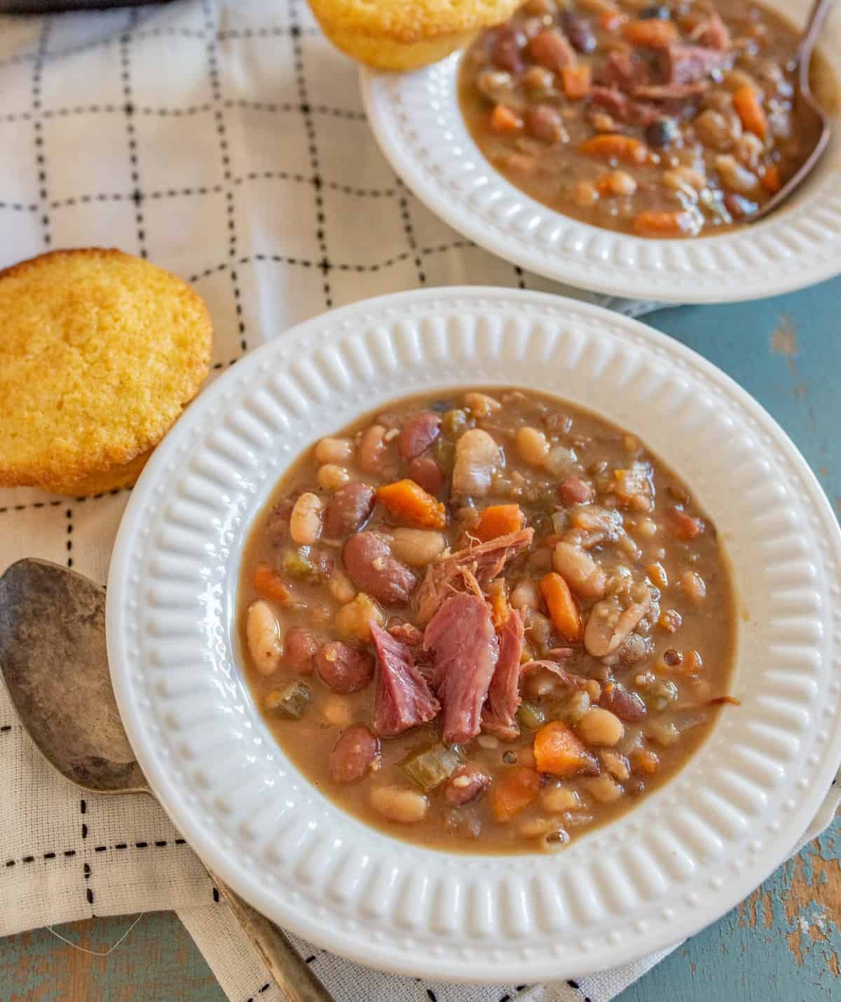 Top View of two white bowls of bean 15 bean soup with a corn muffin on white towel with a spoon.