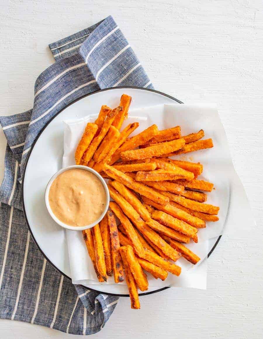 Image of sweet potato fries on a white serving plate with a light red dipping sauce in a round white bowl.