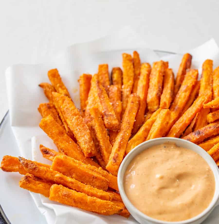 Image of a pile of sweet potato fries on a piece of white parchment paper on a white plate with a small bowl with a light red dipping sauce.