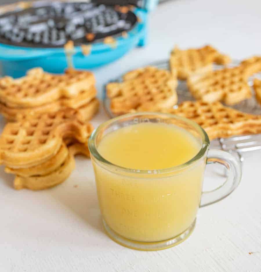 Buttermilk syrup in a small clear glass measuring cup with a blue waffle maker, some eggs, and extra animal-shaped waffles in the background.