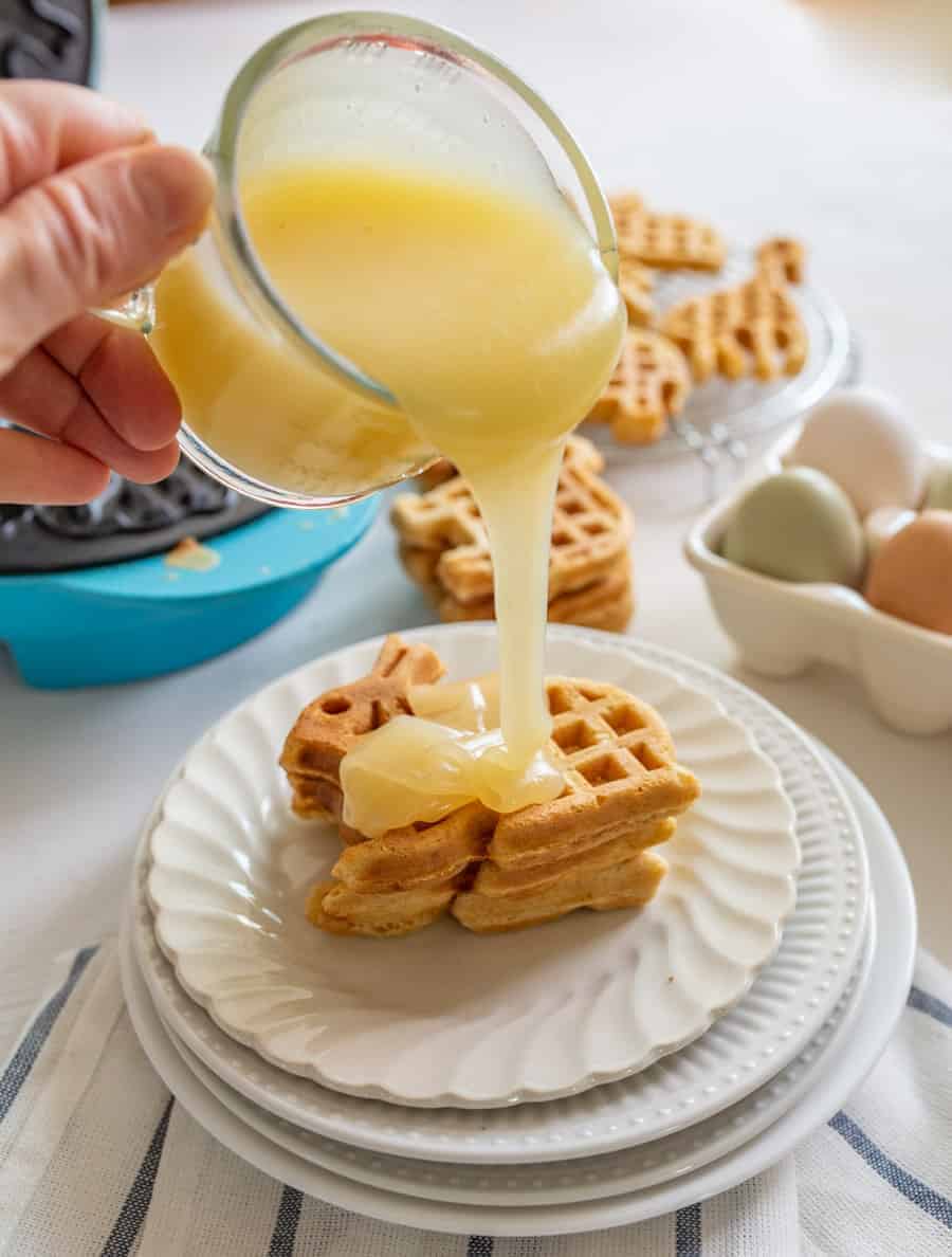 Buttermilk syrup pouring out of a small clear glass measuring cup onto animal-shaped waffles with a blue waffle maker, some eggs, and extra animal-shaped waffles in the background.