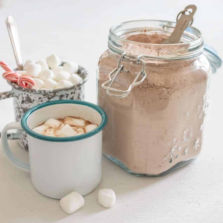Image of a jar clear glass jar filled with homemade hot cocoa mix, a white enamel cup with a blue rim filled with hot cocoa, and a white enamel cup with grey swirls with mini marshmallows and small candy canes.