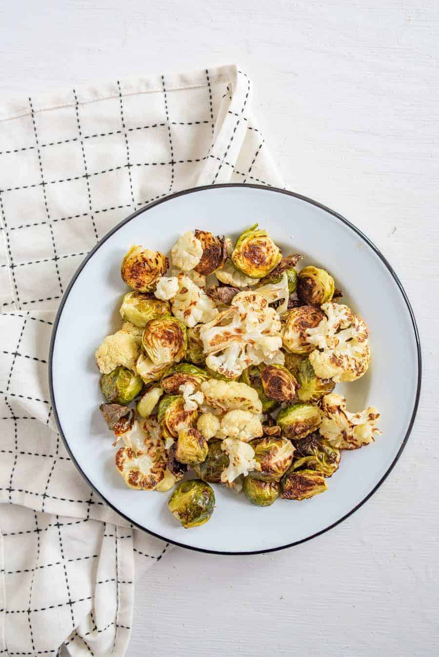 Image of roasted Brussels sprouts and cauliflower on a white plate with a black rim on a white napkin with black accents.