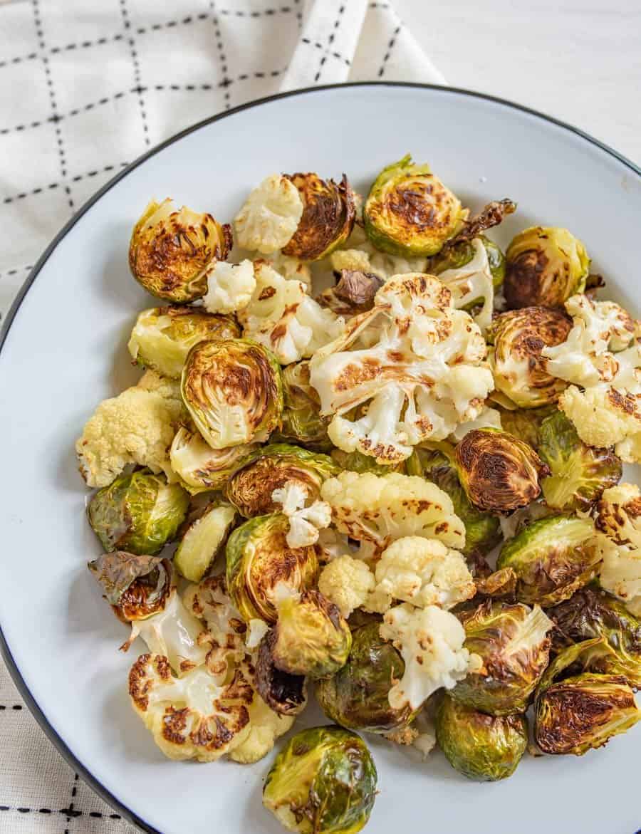 Image of roasted Brussels sprouts and cauliflower on a white plate with a black rim on a white napkin with black accents.