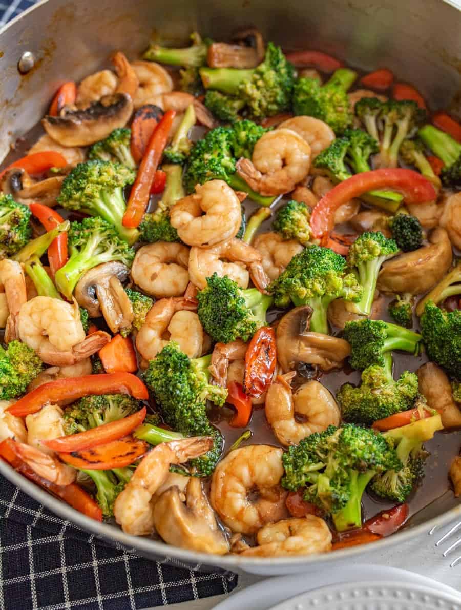 Shrimp stir fry sizzles in a pan with broccoli, red bell peppers, and mushrooms, all tossed in a savory sauce and set on a dark checkered cloth.