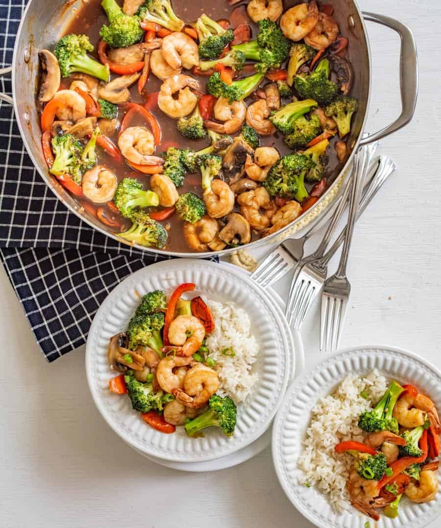 A skillet of shrimp stir fry recipe with broccoli, red peppers, and mushrooms sits beside two plates of white rice topped with the savory dish. Four forks rest on a checkered cloth nearby.