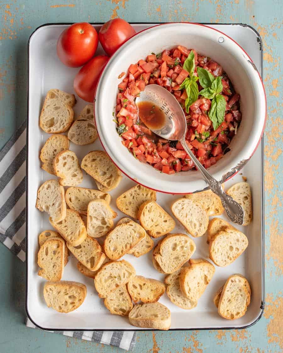 small slices of bread on a white sheet pan with red tomatoes and a white bowl of chopped tomatoes with green basil for summer tomato bruschetta and a silver spoon.