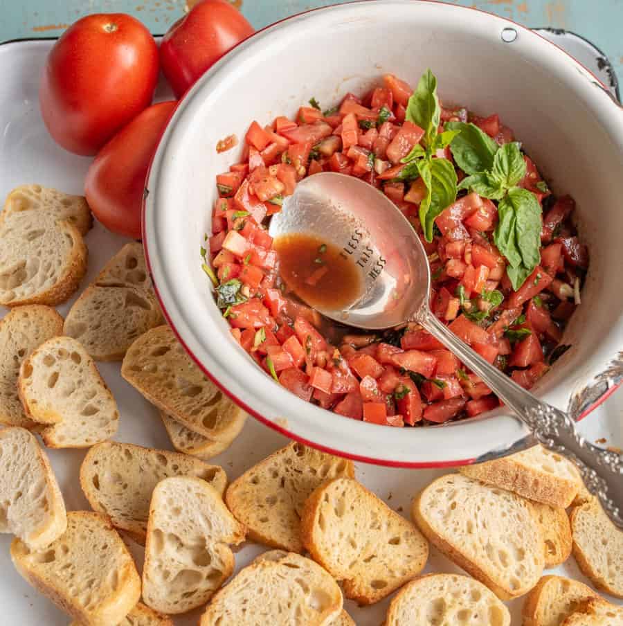 small slices of bread on a white sheet pan with red tomatoes and a white bowl of chopped tomatoes with green basil for summer tomato bruschetta and a silver spoon.