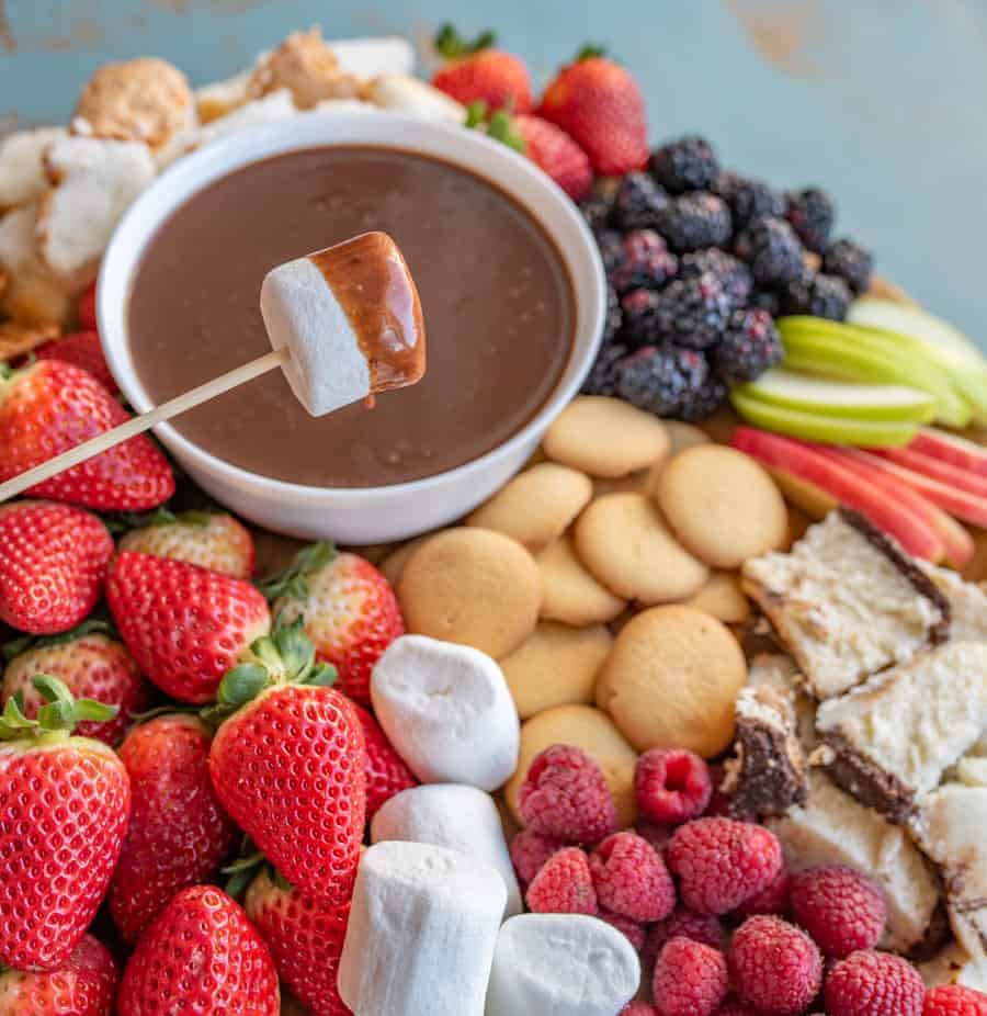 Marshmallow on a wooden skewer that has been dipped in chocolate fondue with things to dip into fondue in the background including strawberries, vanilla wafers, apple slices, blackberries, pound cake, raspberries, and angel food cake.