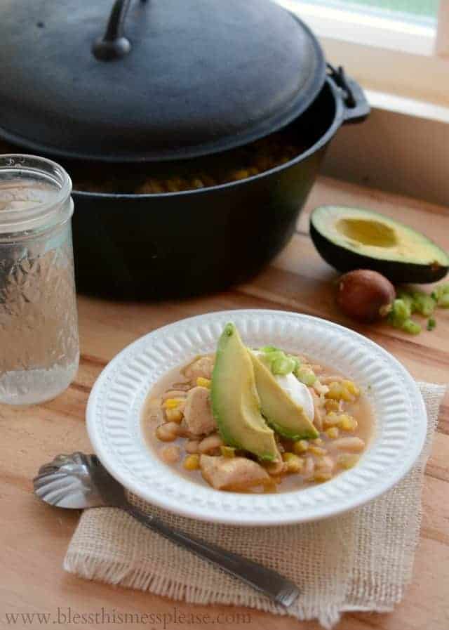 Image of a white bowl with white chicken enchilada soup garnished with two avocado slices, some sour cream, and sliced green onions.