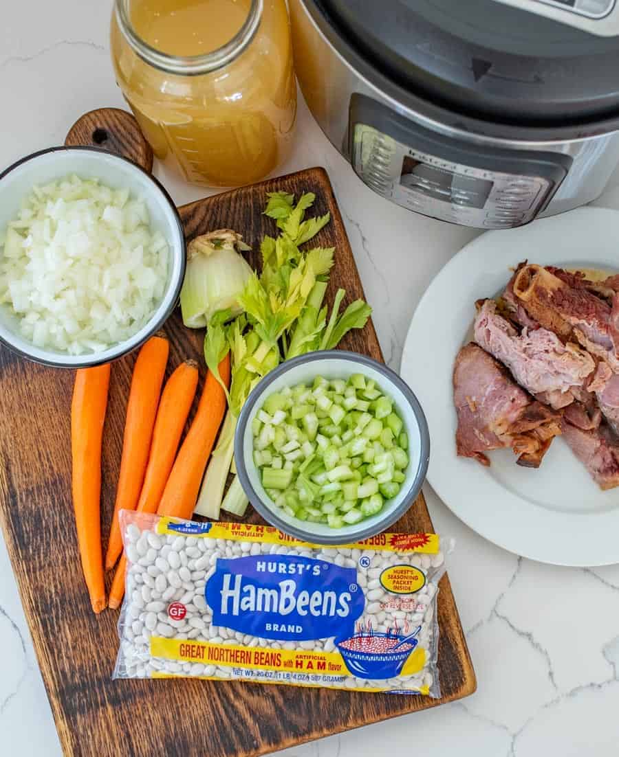 Ingredients for ham and bean soup are laid out on a cutting board: packaged white beans, chopped onions, celery, whole carrots, and garlic. Nearby sit a plate with ham hocks, a bowl of broth, and an Instant Pot ready for action.