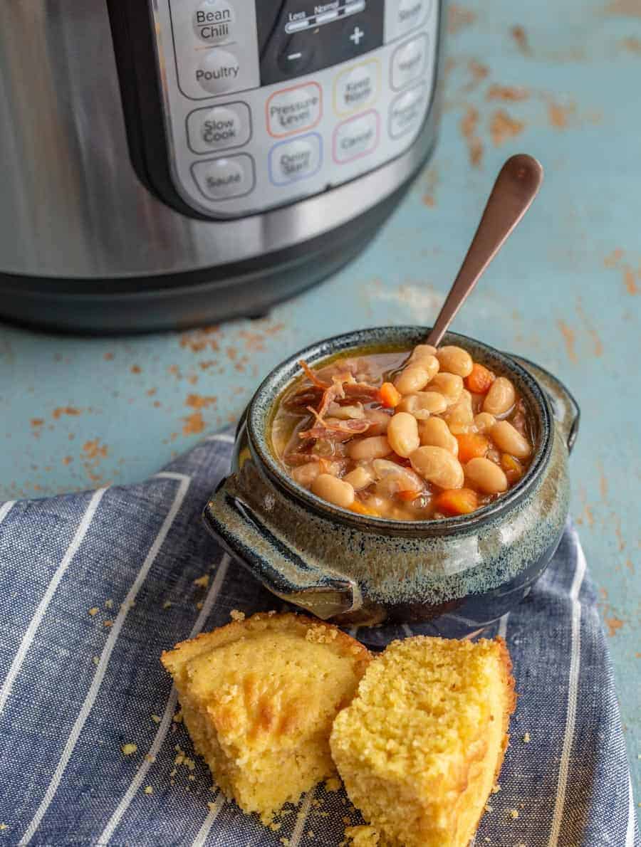 A bowl of bean chili with a spoon rests on a blue-striped cloth beside cornbread pieces, reminiscent of a hearty ham and bean soup. In the background, the pressure cooker with labeled buttons stands ready for its next culinary adventure.