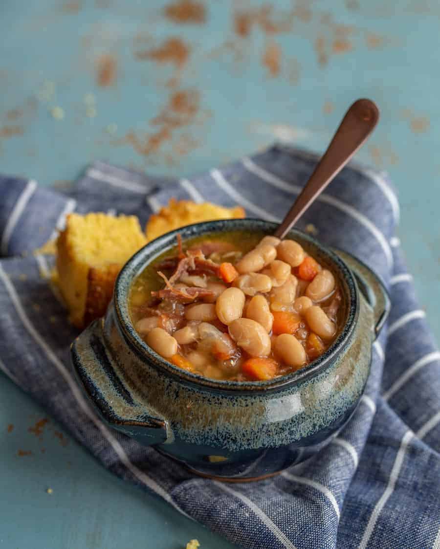 A rustic blue ceramic bowl brimming with a hearty ham and bean soup recipe, featuring succulent chunks of meat and diced carrots, is served with a spoon. The scene is completed by pieces of cornbread resting on a striped cloth atop the textured blue surface.