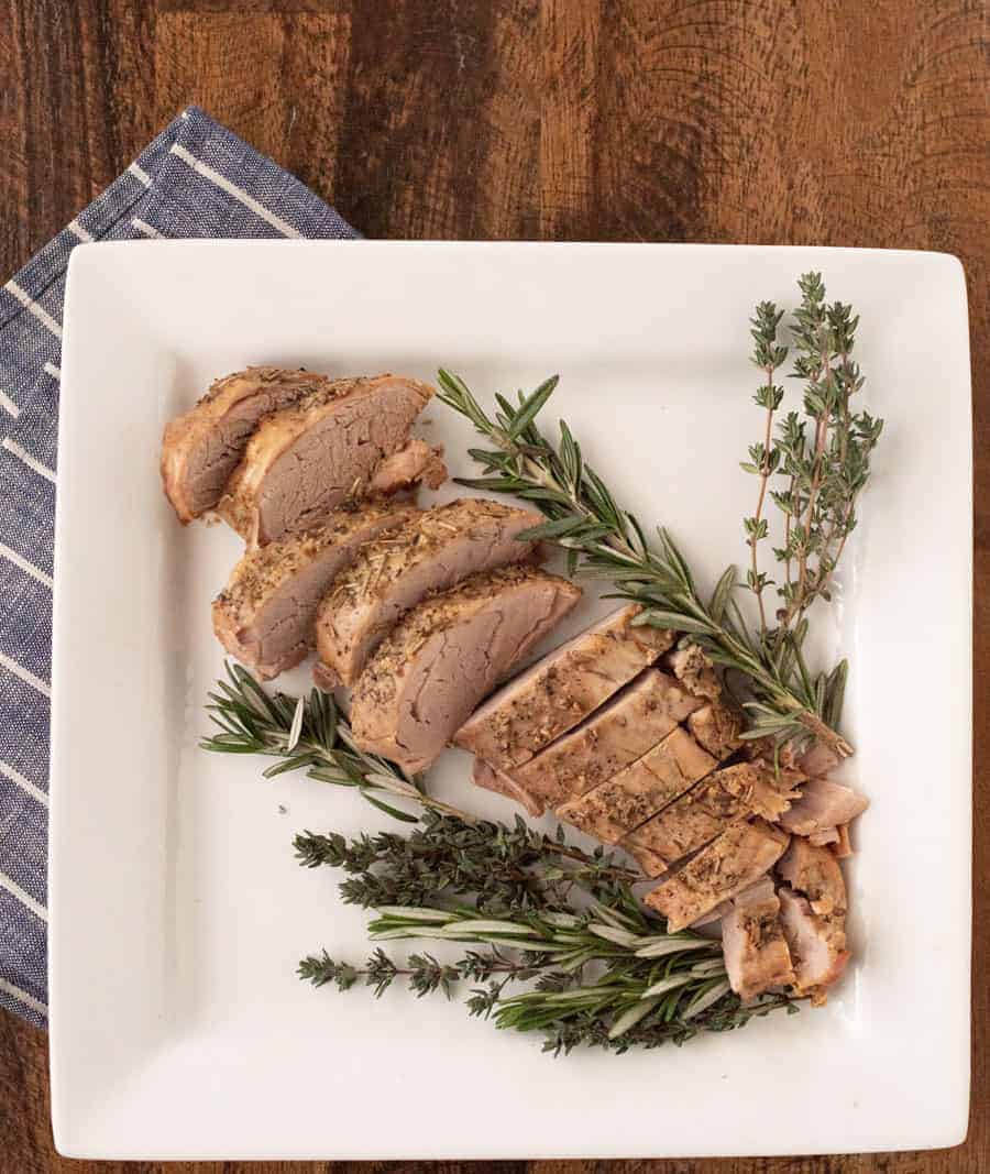 Crock pot cooked pork loin sitting on a white plate, sliced, with fresh herbs around it.