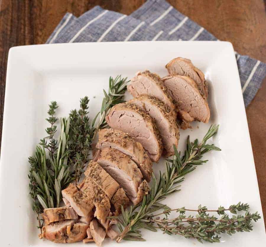 A plate of crock pot cooked pork loin, sliced, with fresh herbs around it.