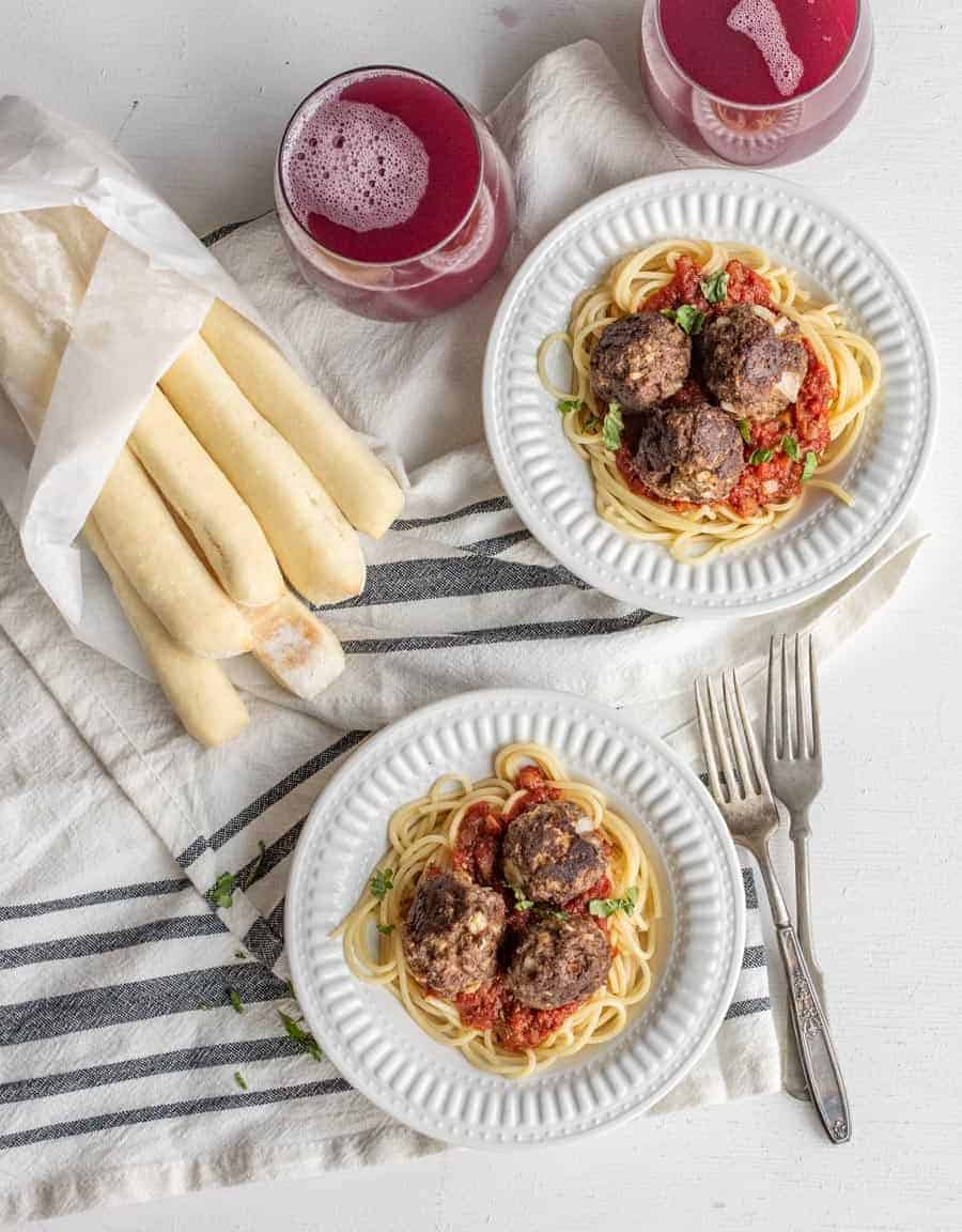 Homemade meatballs on top of a plate of spaghetti with marinara sauce with grape juice and bread sticks in the background.
