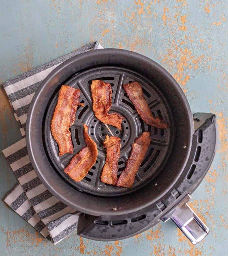 Crispy bacon in Air Fryer, perfectly cooked strips rest in a basket atop a black and white striped napkin on a distressed blue surface.