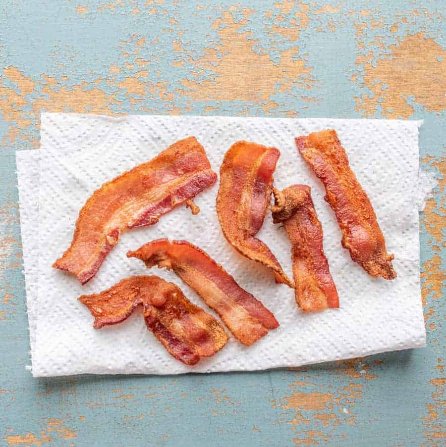 Six crispy strips of bacon, cooked to perfection in the air fryer, rest on a white paper towel atop a light blue surface with hints of exposed wood peeking through.