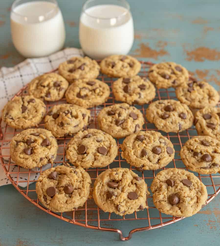 A batch of chocolate chip cookies cooling on a wire rack, with two glasses of milk in the background. The cookies are arranged neatly on the rack next to a few delicious peanut butter oatmeal cookies, and the scene is set on a rustic, turquoise-painted surface.