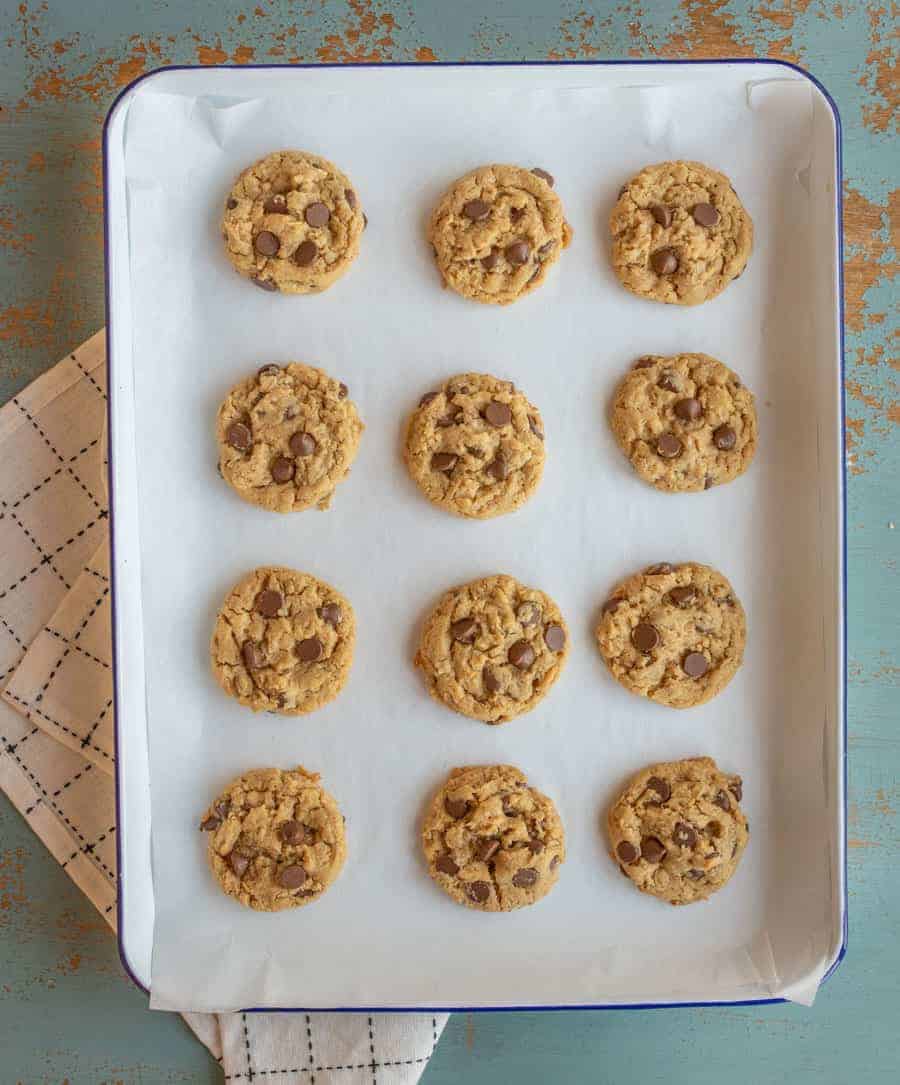Twelve warm, chocolate chip cookies sit in three rows on a parchment-lined baking tray. On the blue surface beneath, a black and white checkered cloth peeks out, hinting at delicious peanut butter oatmeal cookies waiting to be baked next.